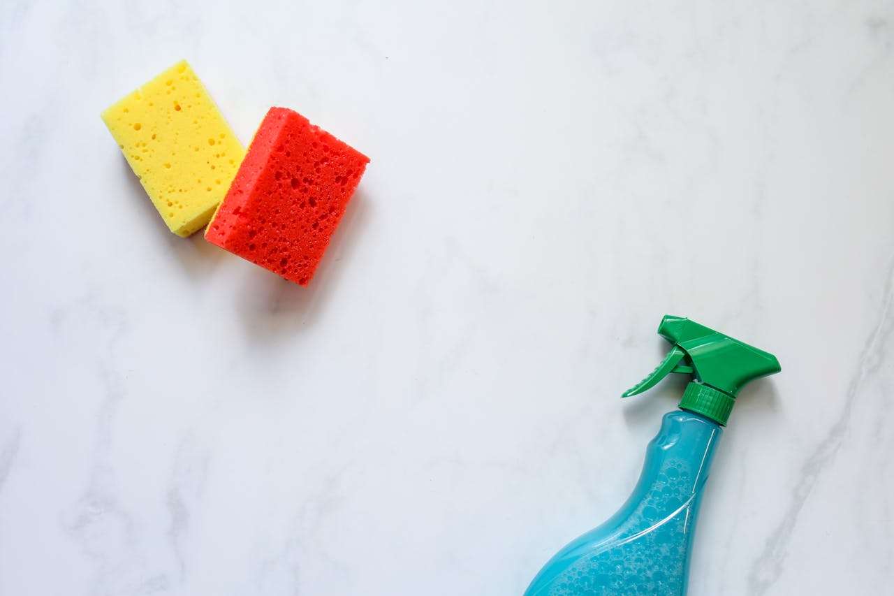 Red and yellow sponges with a blue spray bottle on a marble background, offering cleaning themes.